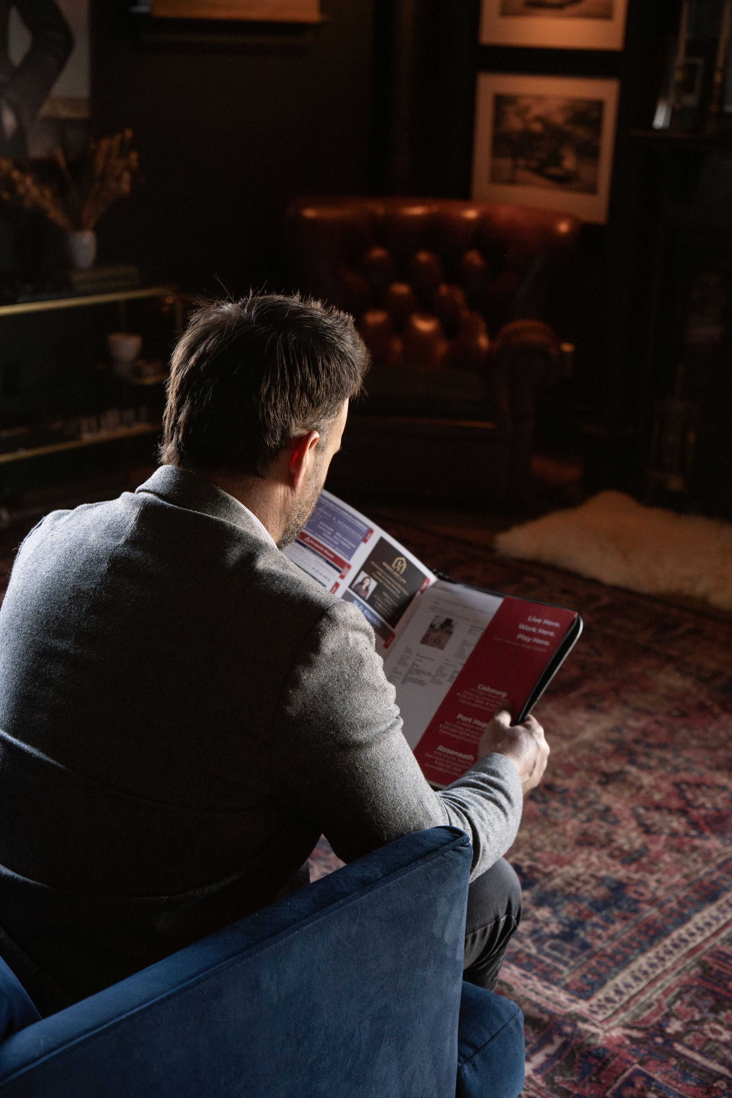Someone reads a book while relaxing in a cozy armchair on a patterned rug in a dimly lit room with framed artwork.