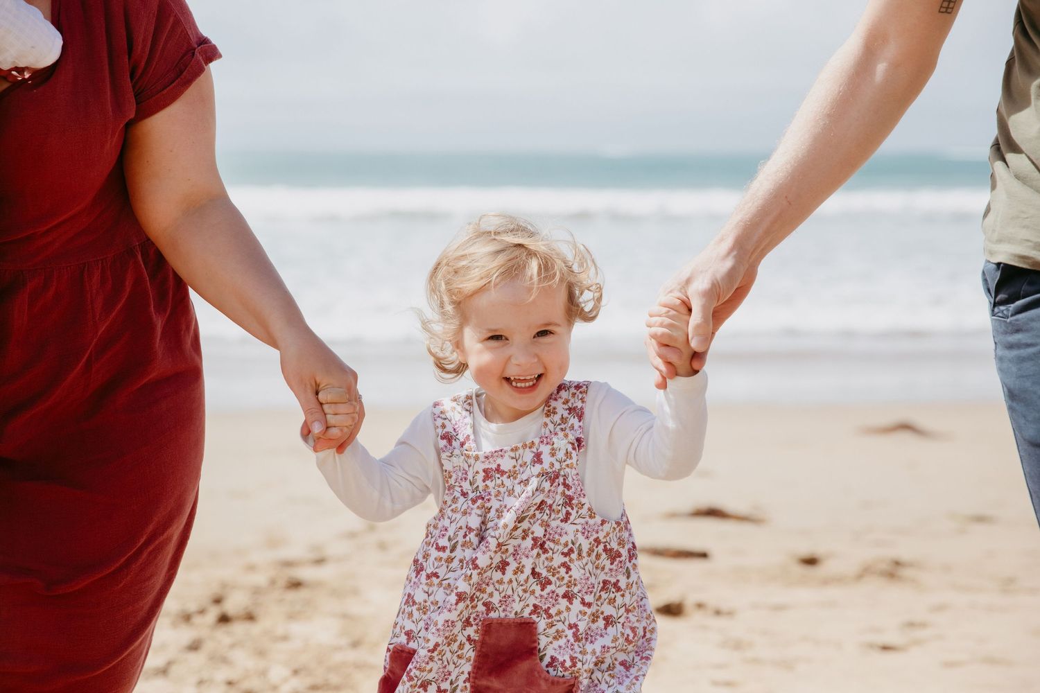 A joyful child in a floral dress walks along the beach while holding hands with adults.
