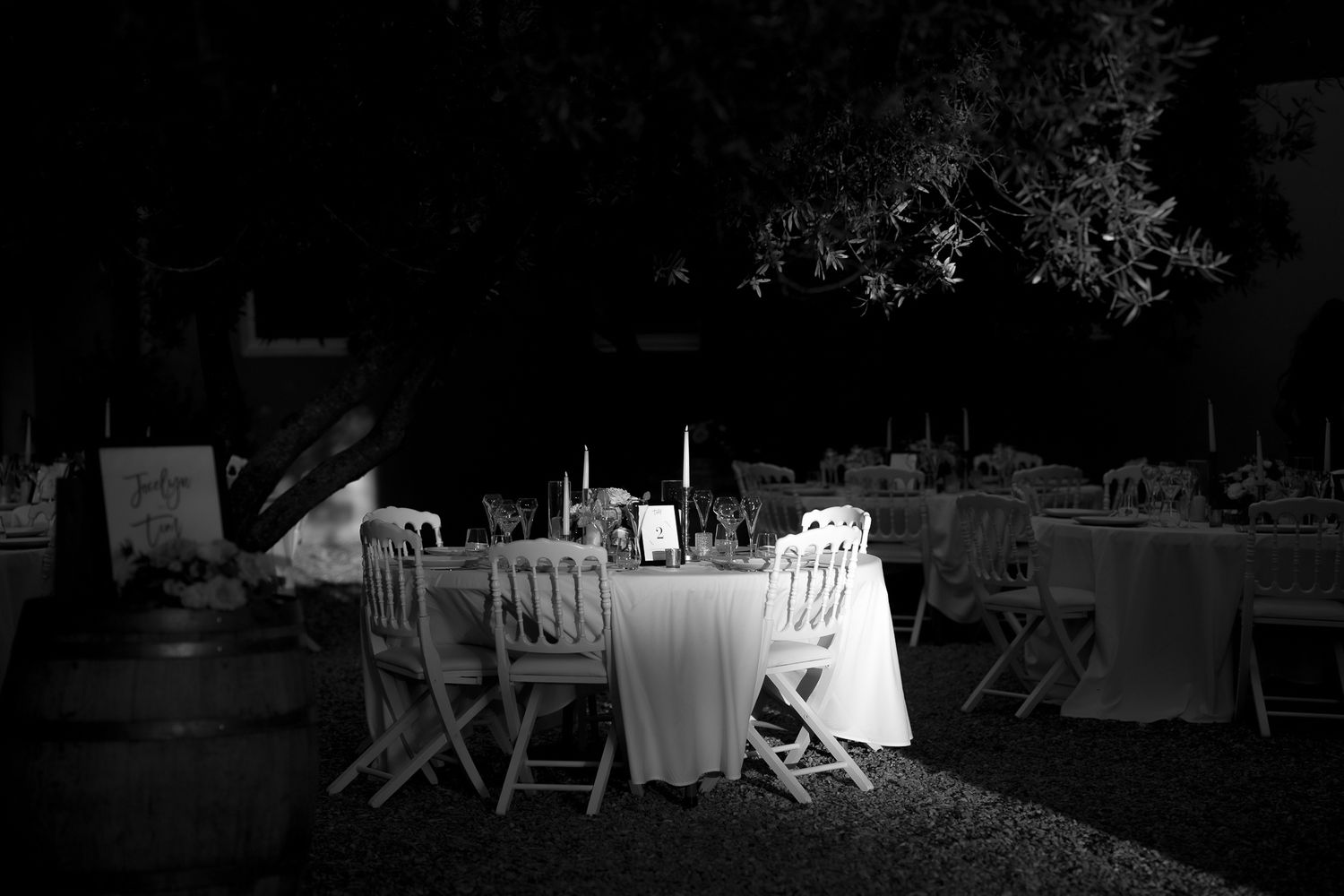 A black and white photograph of an empty outdoor dining table with white tablecloth and chairs illuminated in darkness.