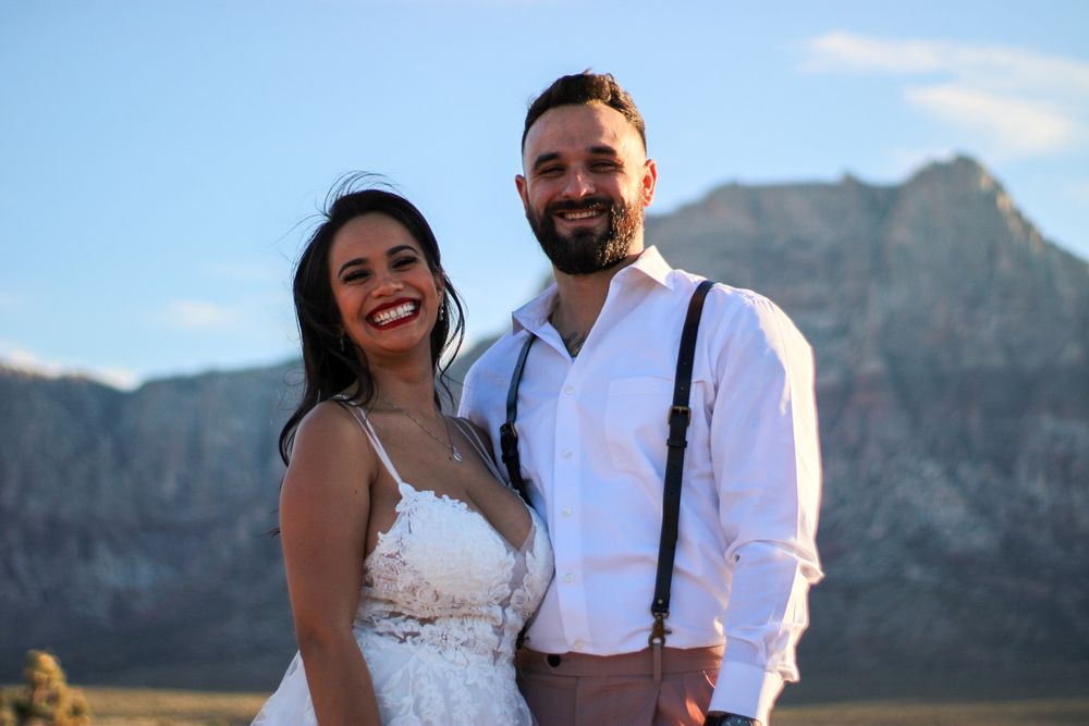 A smiling couple in wedding attire poses outdoors with a scenic mountain backdrop at golden hour.