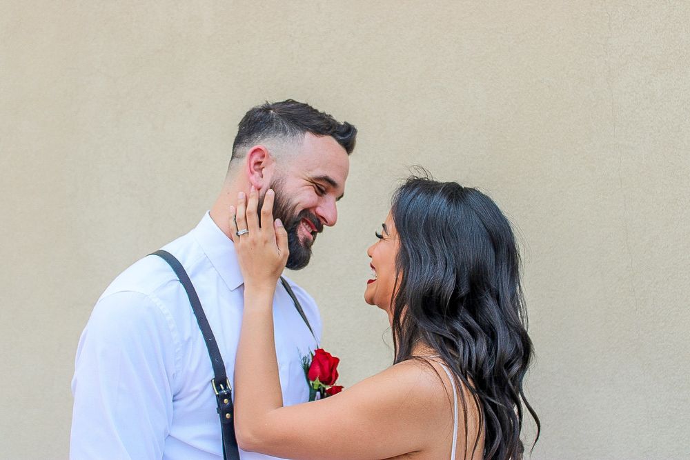 Bride touching bearded grooms face while holding red rose against neutral wall background.
