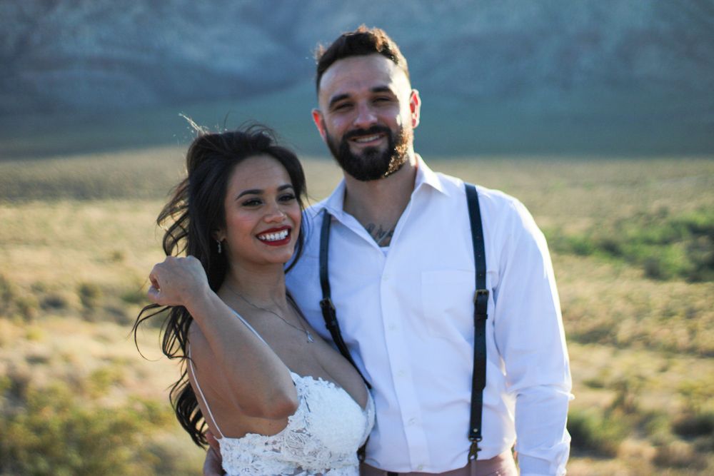 Happy couple posing outdoors in desert landscape, bride in lace dress and groom in white shirt with suspenders.