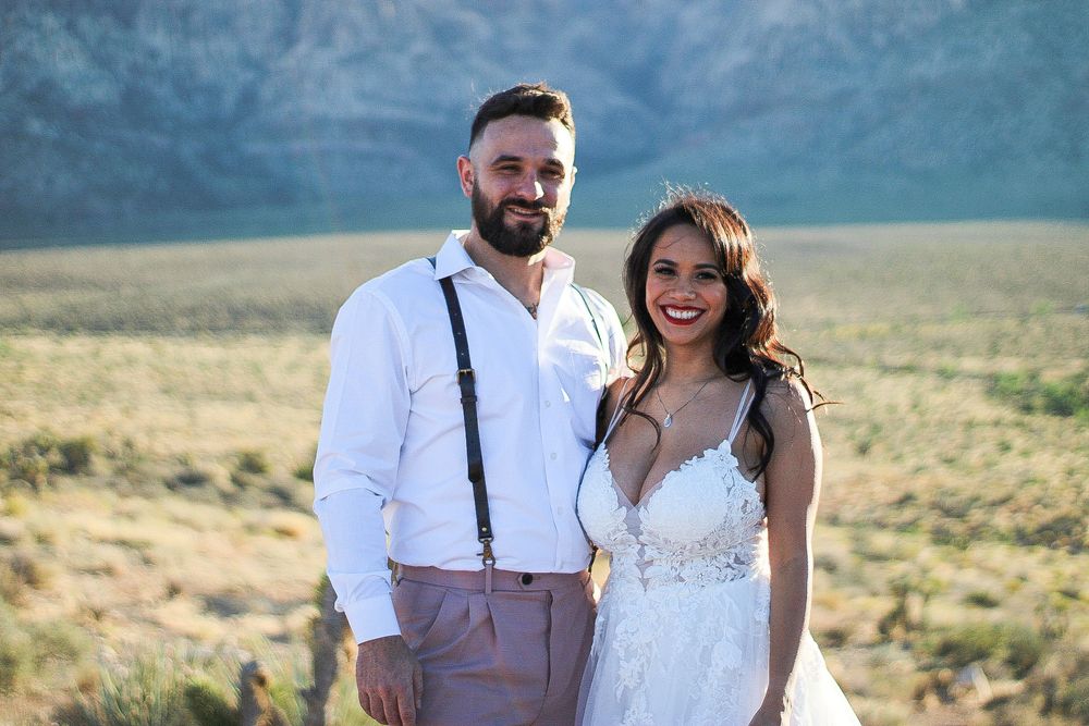 Smiling bride and groom posing together in desert landscape with mountains in background.