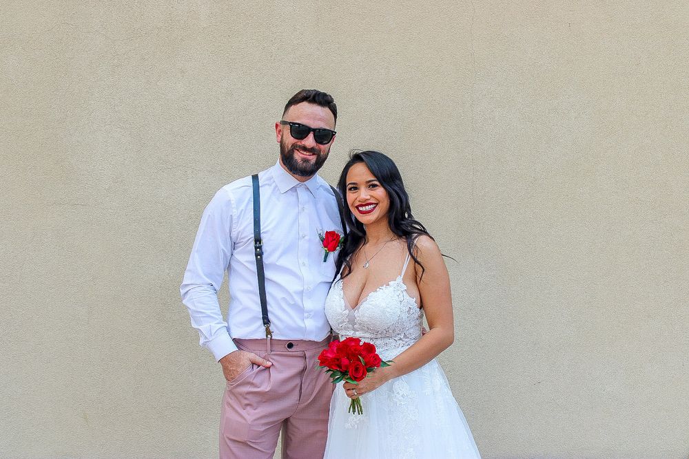 Happy bride holding red roses bouquet standing with groom wearing suspenders against beige wall.