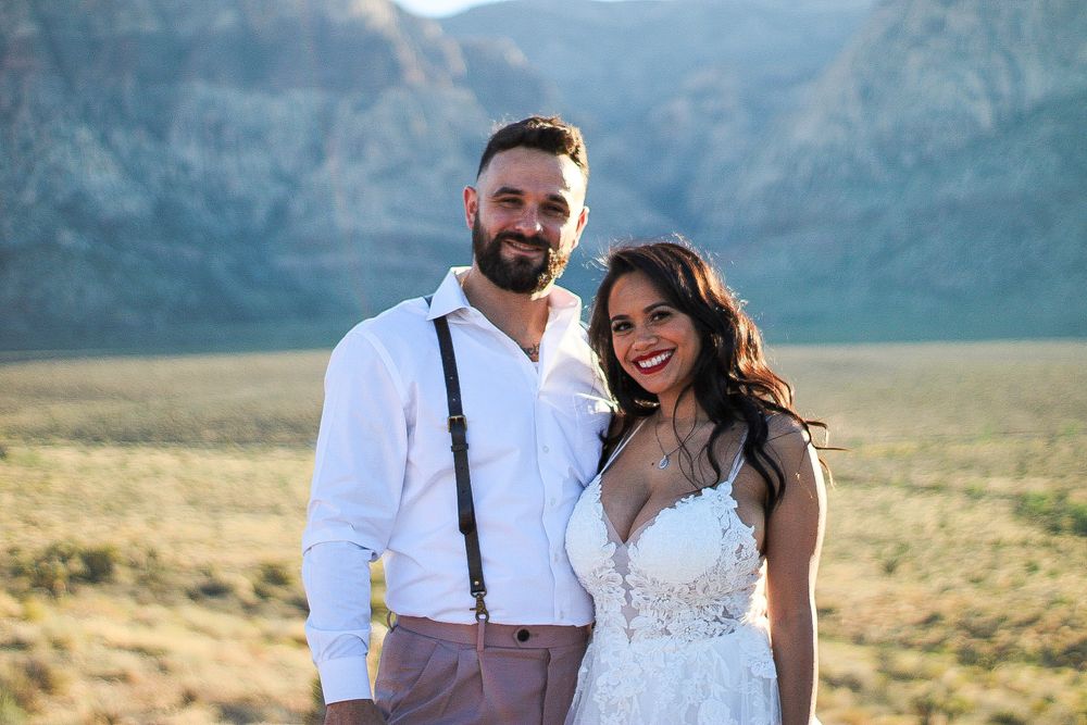 Bride and groom smiling outdoors with scenic desert mountain landscape glowing in sunlight.