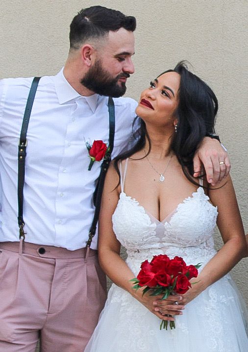 Bride and groom in wedding attire, she holds red roses, he wears suspenders with a boutonniere, gazing at each other.
