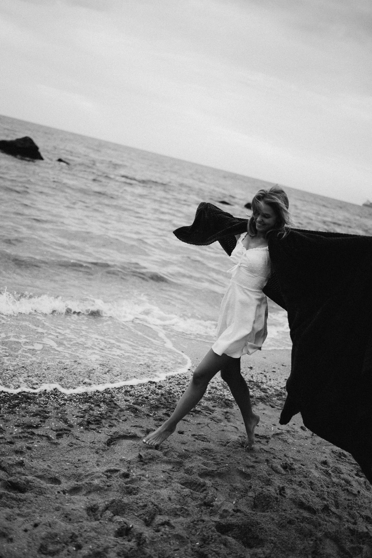 Woman in white dress walking on a beach with a black cape blowing in the wind.