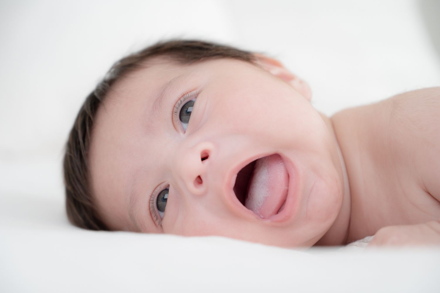 Baby with bright eyes and open mouth lies on white bedding.