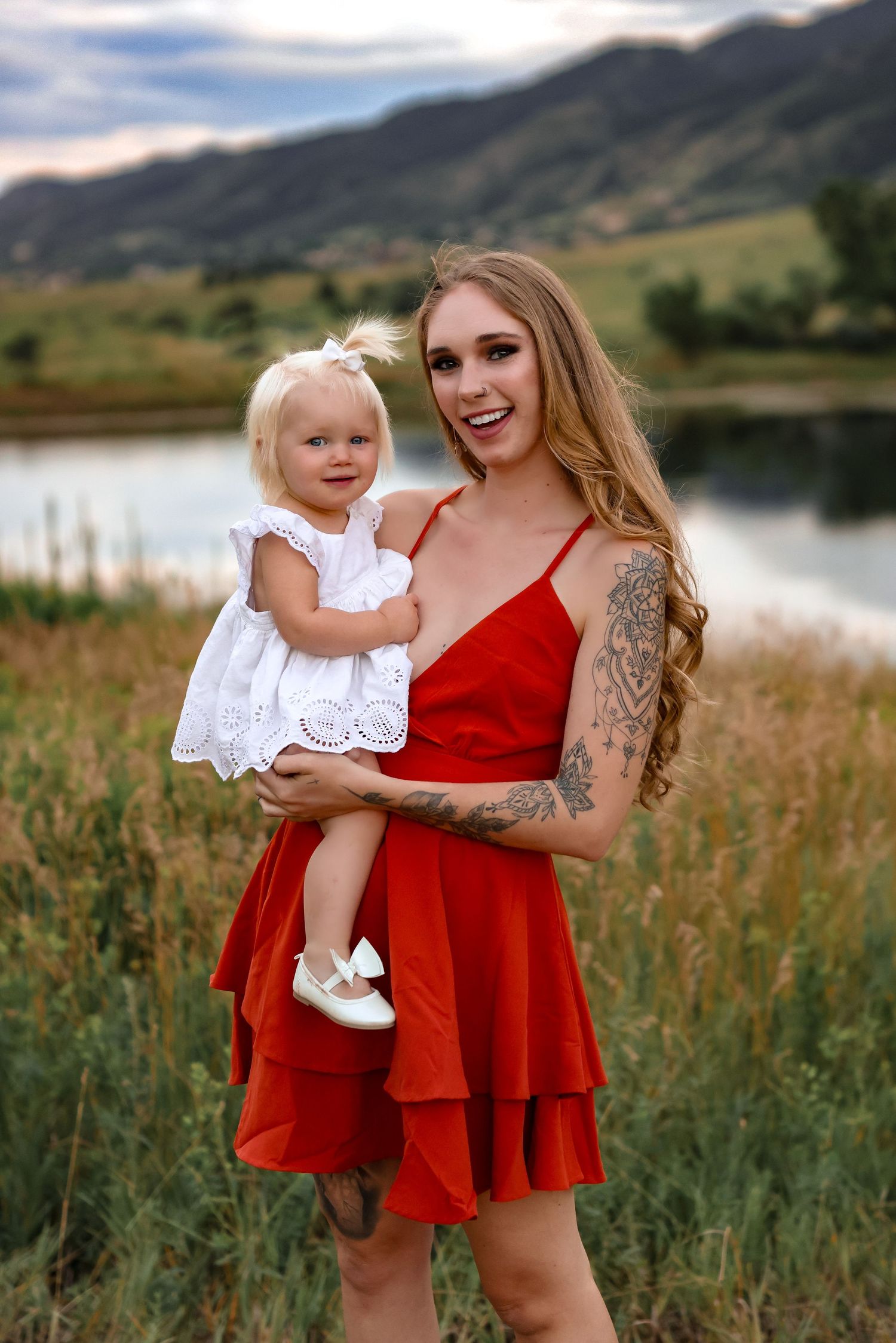A tender moment captured by a lake with mountains in the background, featuring summer dresses in red and white.