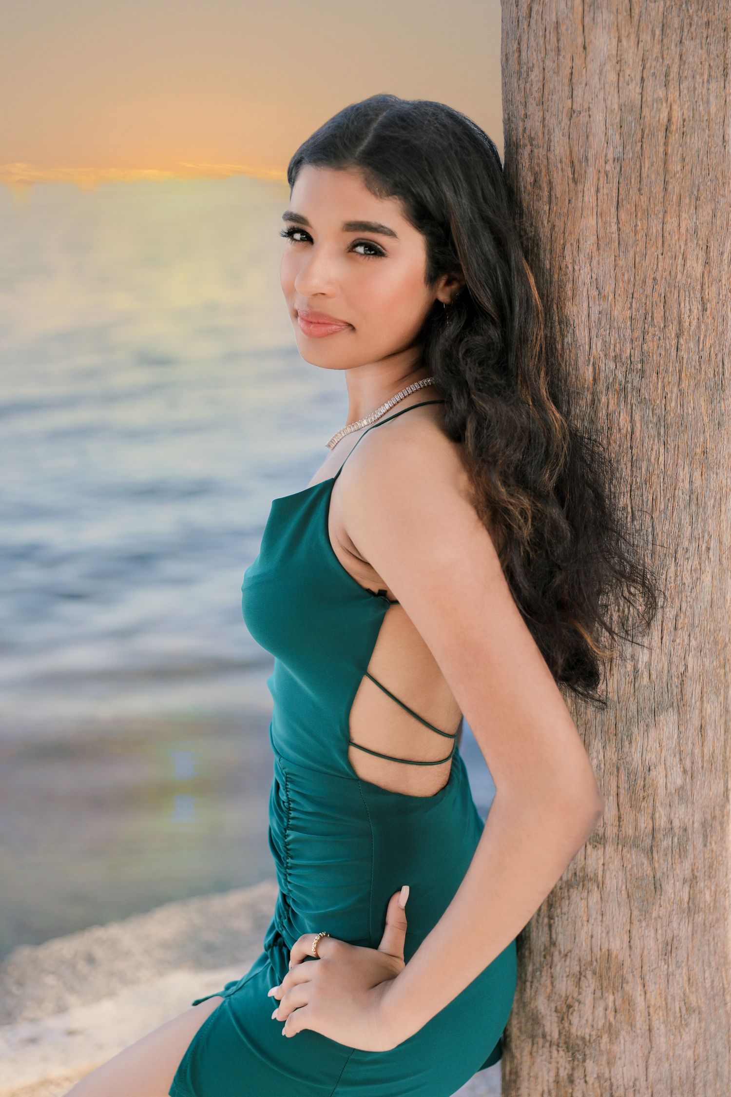 Woman in emerald green dress poses against wooden post at sunset by the ocean.