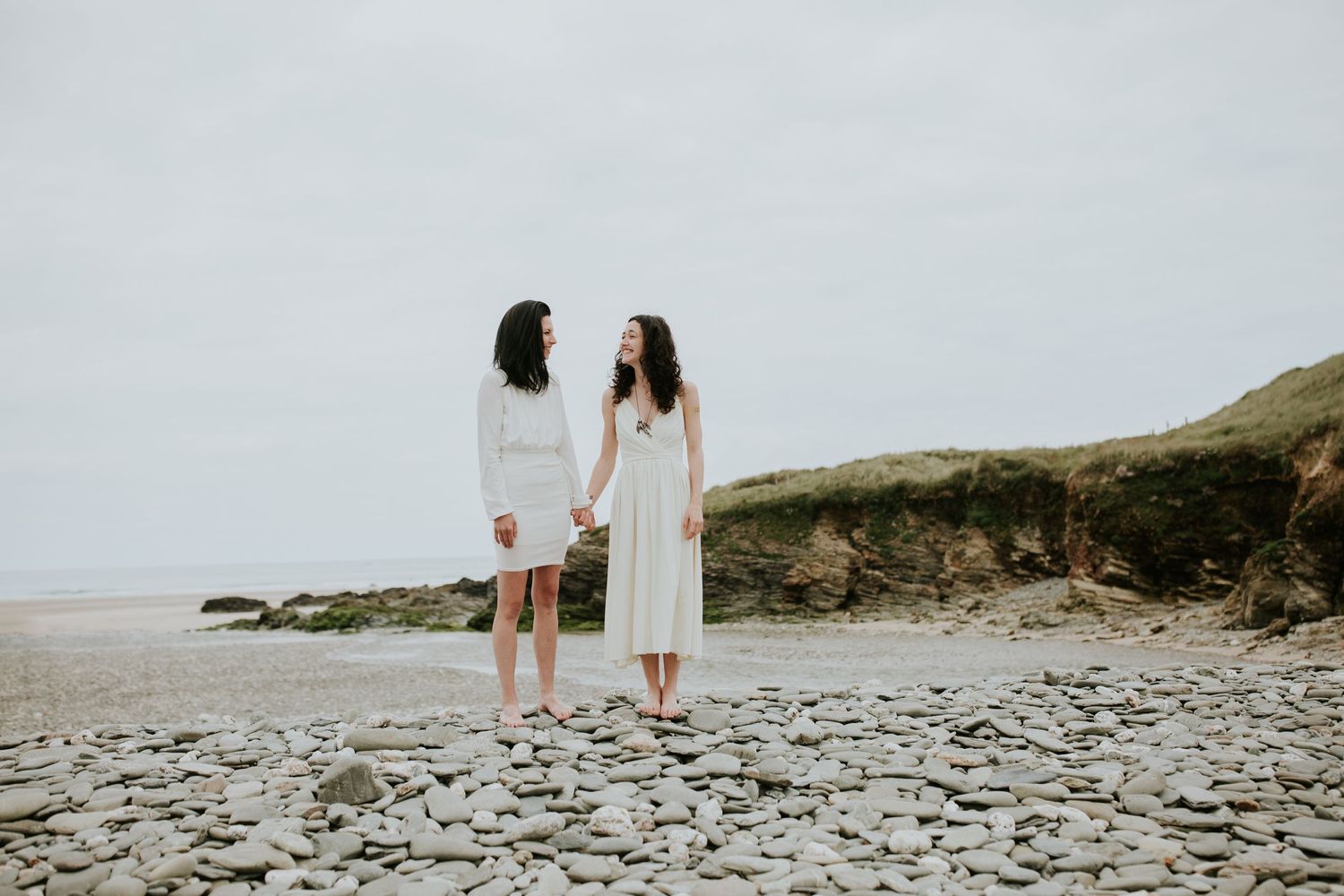 Two people in white dresses standing together on a rocky beach with cliffs in the background on an overcast day.