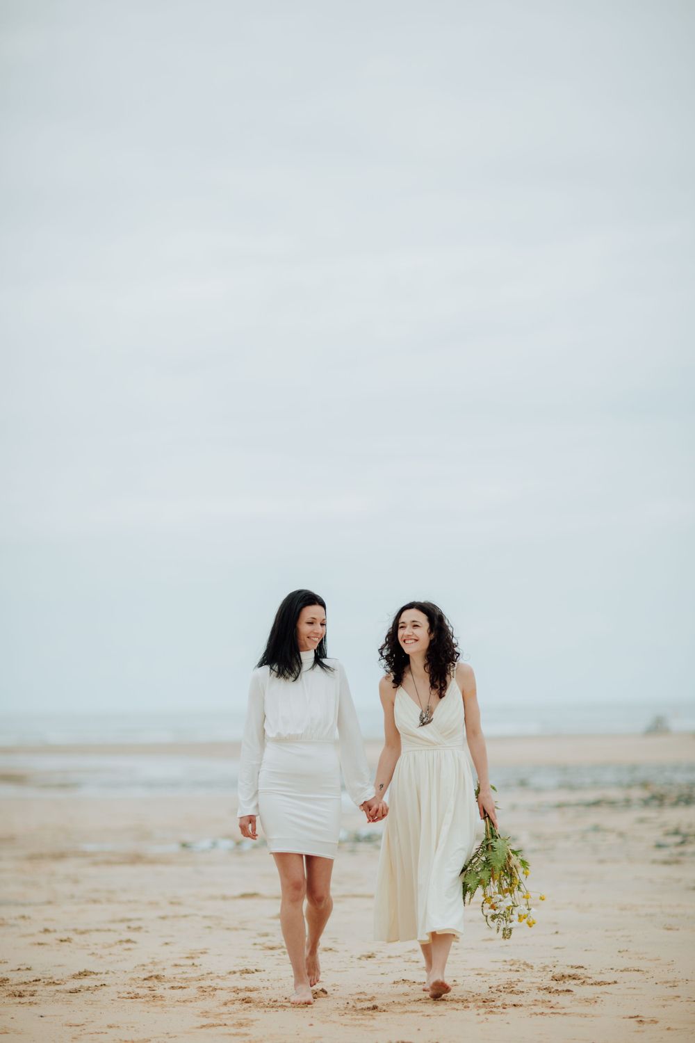 Two people in white dresses walking hand in hand along a scenic beach.