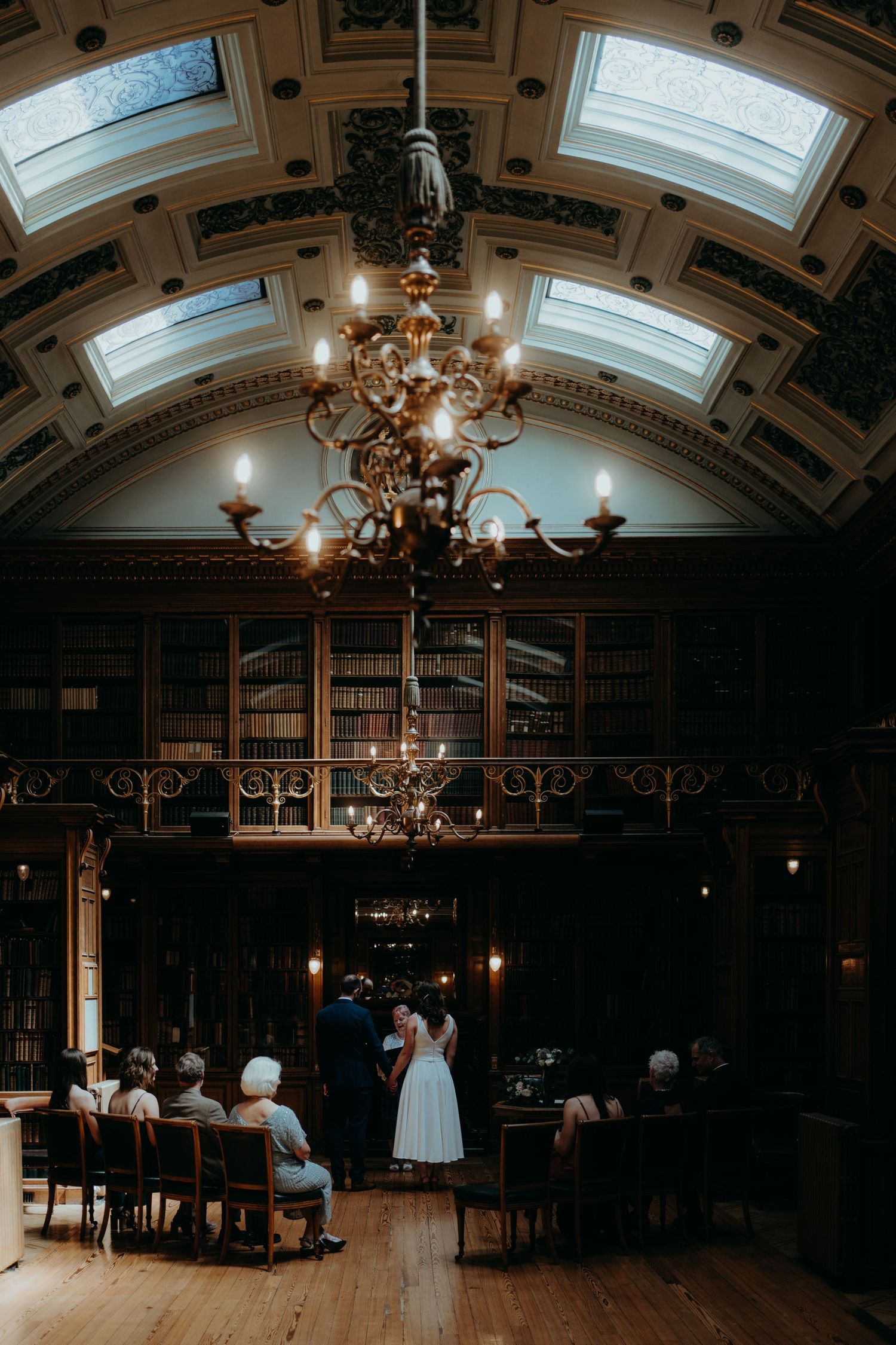 Wedding ceremony in an elegant room with ornate ceiling and fireplace surrounded by seated guests.