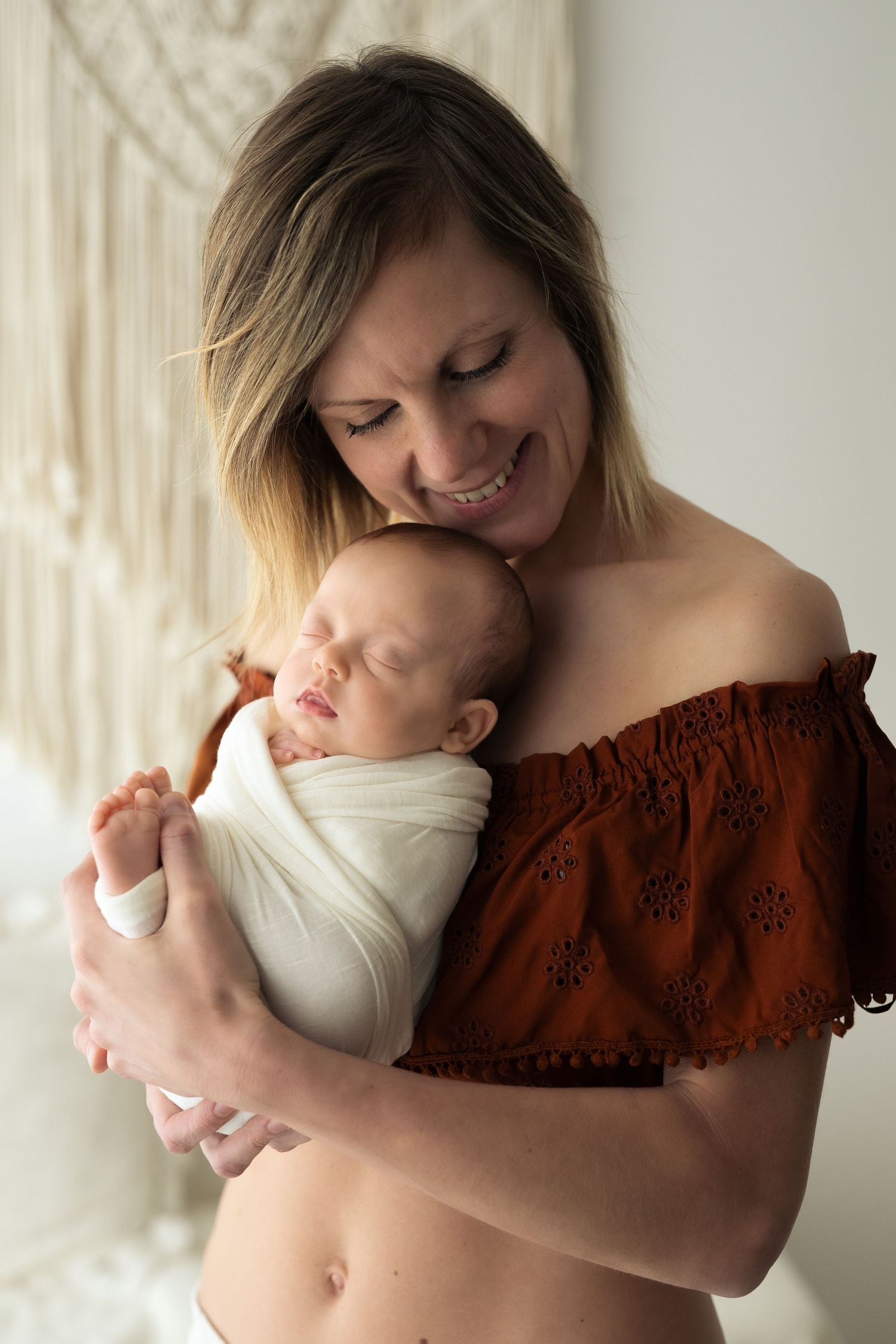 Bellissima mamma sorride mentre tiene sul petto la sua neonata completamente avvolta di una fasciatura che la contiene, in un set romantico.