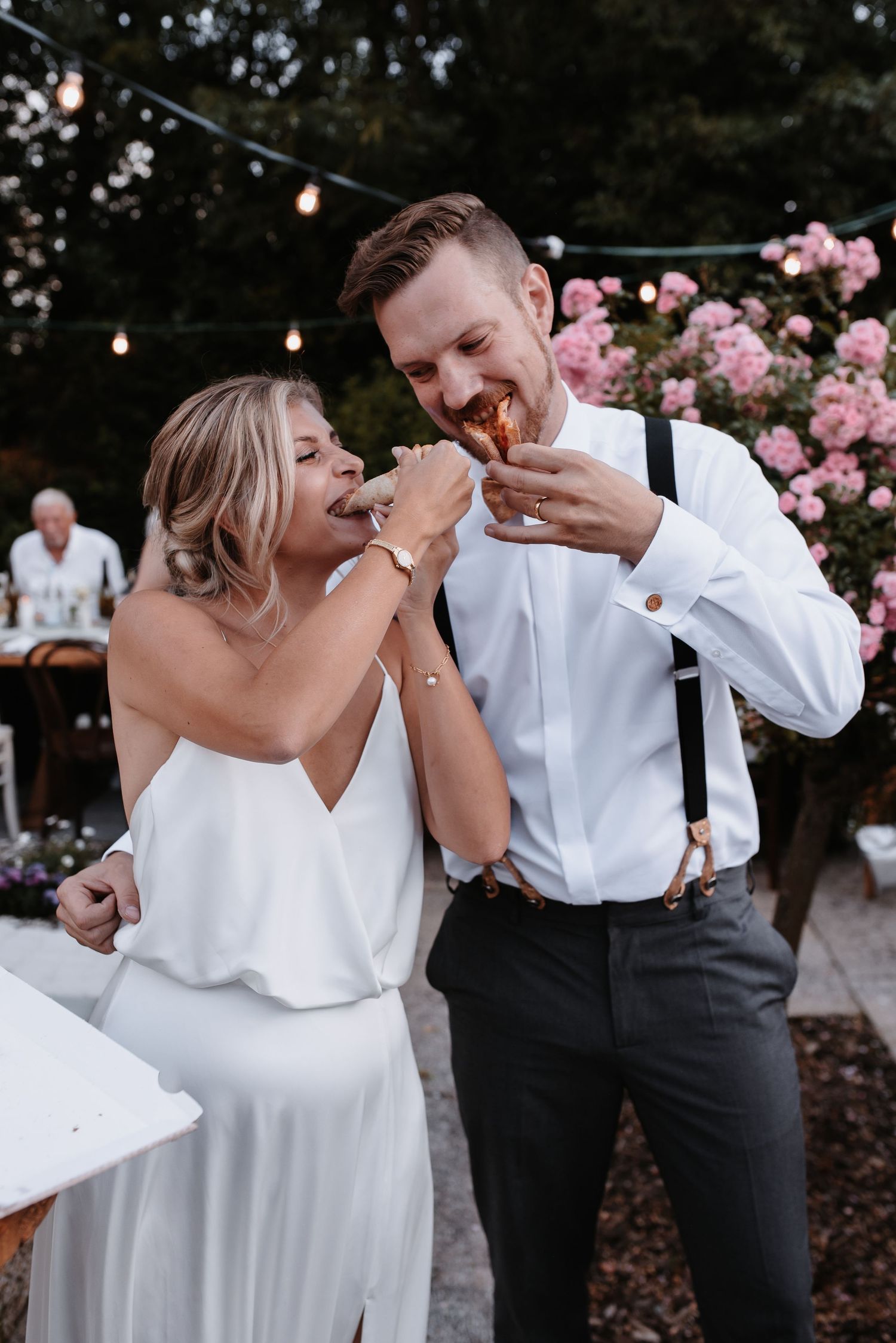 A candid wedding moment shows a couple sharing dessert and laughing together outdoors.