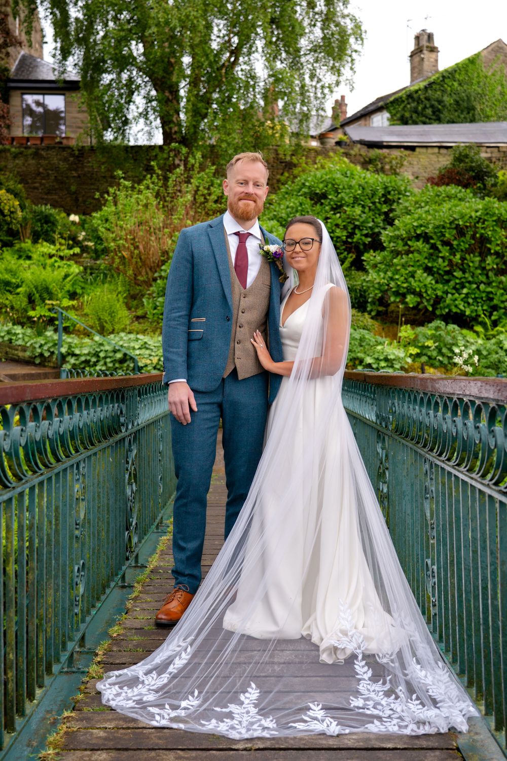 Newlywed bride and groom stood on an iron bridge over the river in Bollington, Cheshire with her veil spread across the bridge.