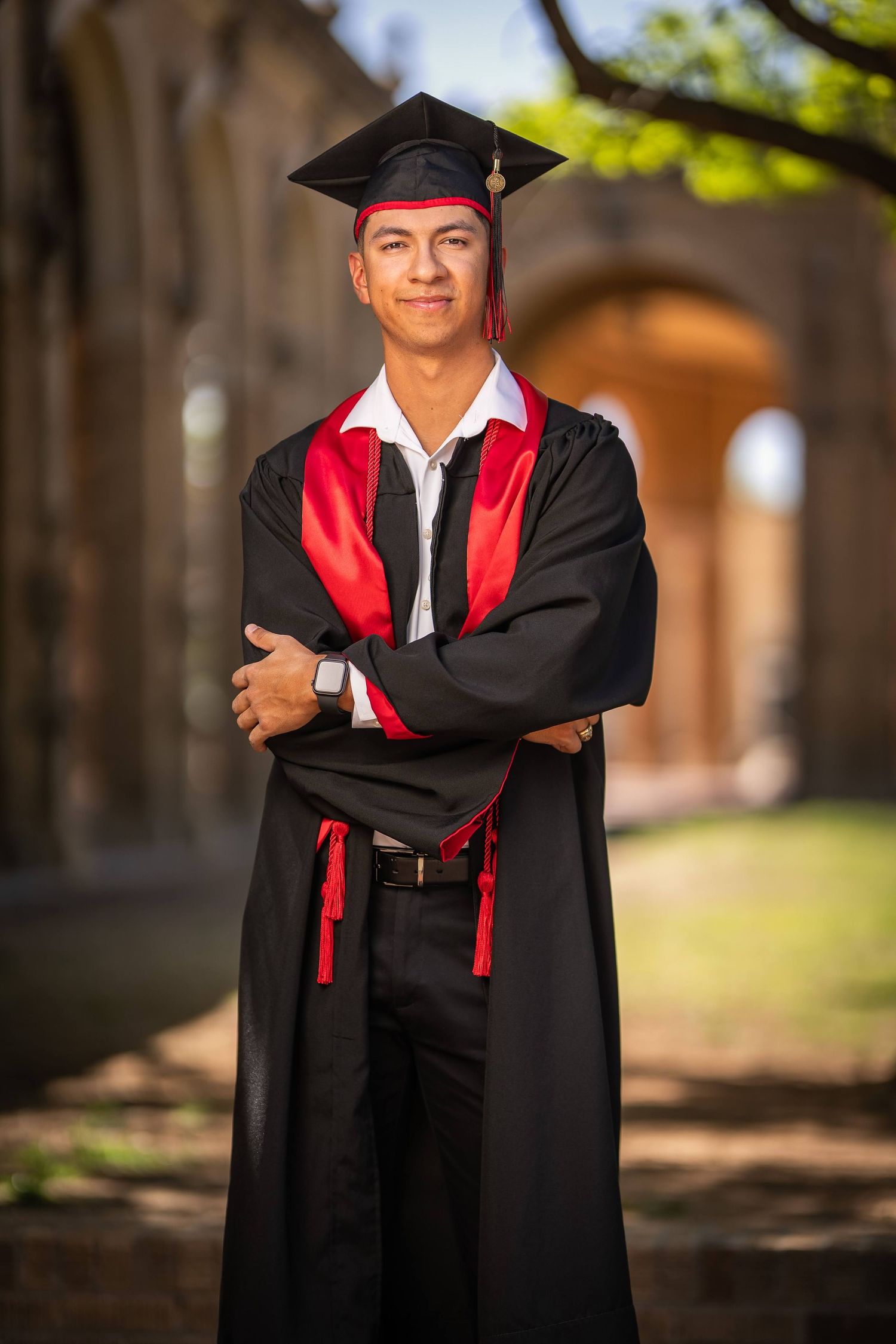 Graduate in traditional black and red academic regalia poses outdoors near stone architecture.