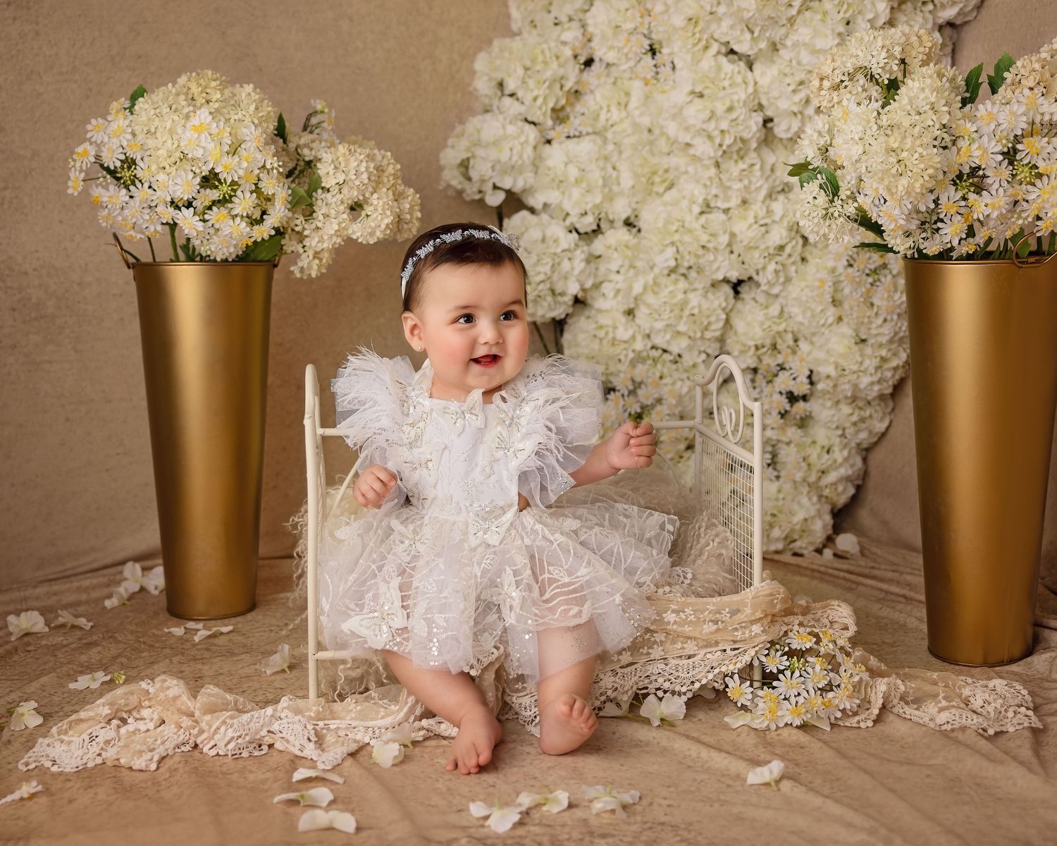 Baby in white lace dress surrounded by elegant floral arrangements in gold vases.