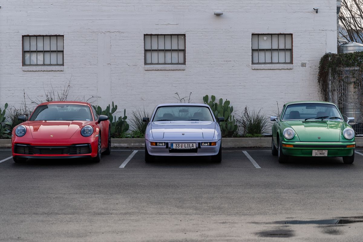 Three sports cars - a red, silver, and green - parked in a row against a white wall with windows.