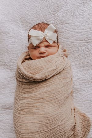 Newborn baby wrapped in beige swaddle with white bow against textured white background.
