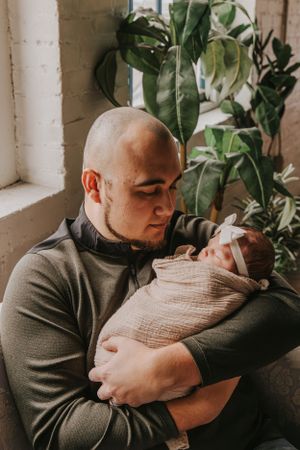 Parent holding swaddled newborn near window with tropical plants.