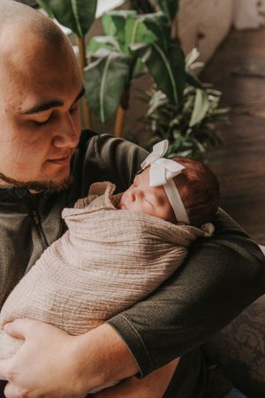 Intimate portrait of parent with swaddled newborn by sunlit window.