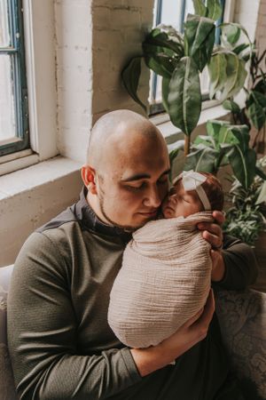 Parent in grey sweater holding swaddled baby near window with plants.
