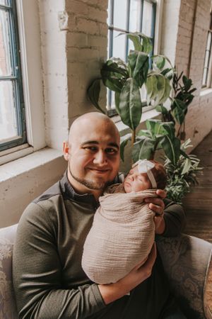 A tender moment between parent and baby wrapped in beige swaddle blanket near indoor plants and windows.