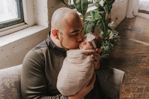 Parent and swaddled infant sharing intimate bonding moment by window with houseplants.
