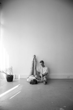 Artistic black and white photo of two people in relaxed poses against white wall with plant.