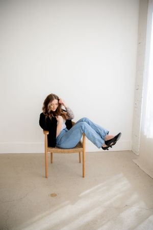 Casual lifestyle portrait on wooden chair against white wall with dramatic shadows.