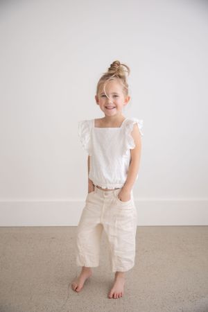 Young child wearing all white outfit with ruffled sleeves and cropped pants poses barefoot against neutral wall.