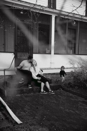 Black and white image of family relaxing on porch while child plays in background.