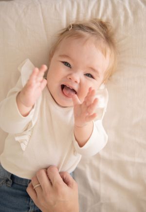 Baby wearing cream sweater laughs joyfully while lying on white bedding.