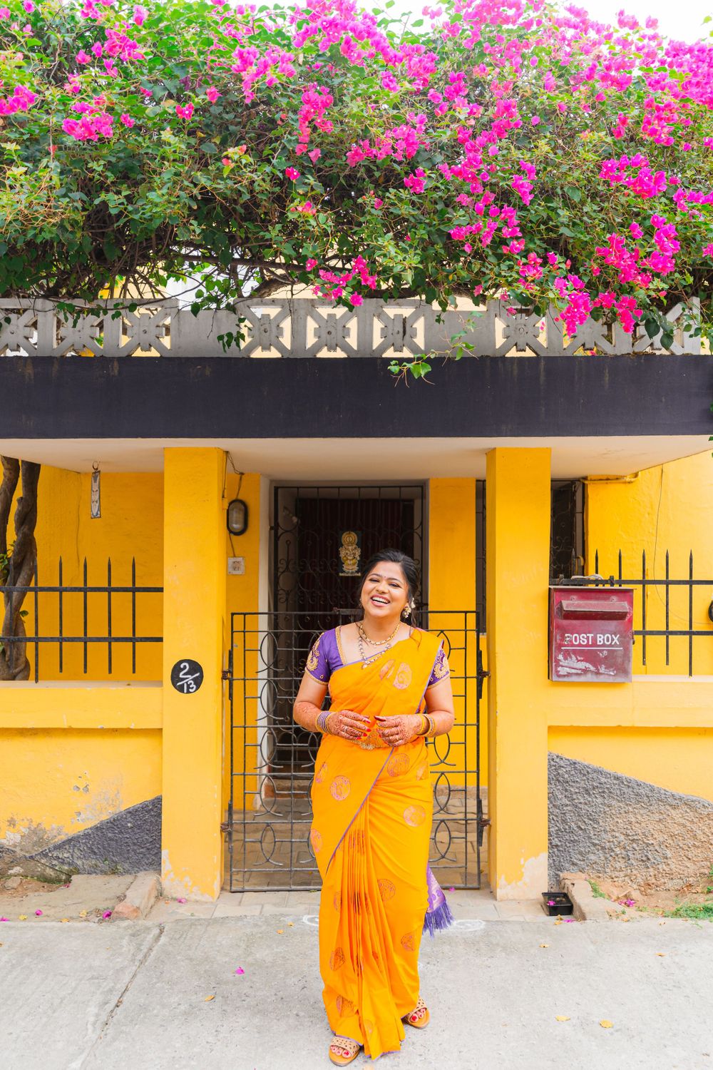 Woman in orange sari stands in front of yellow building with pink bougainvillea flowers.
