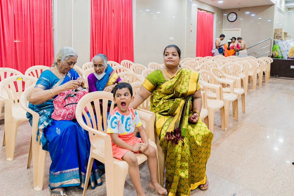 Wedding guests seated in plastic chairs in a hall with red curtains.