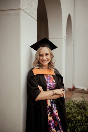 A graduate poses in academic regalia against white arched architecture.