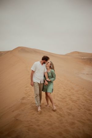 A romantic couple standing together in sand dunes against an overcast desert sky.
