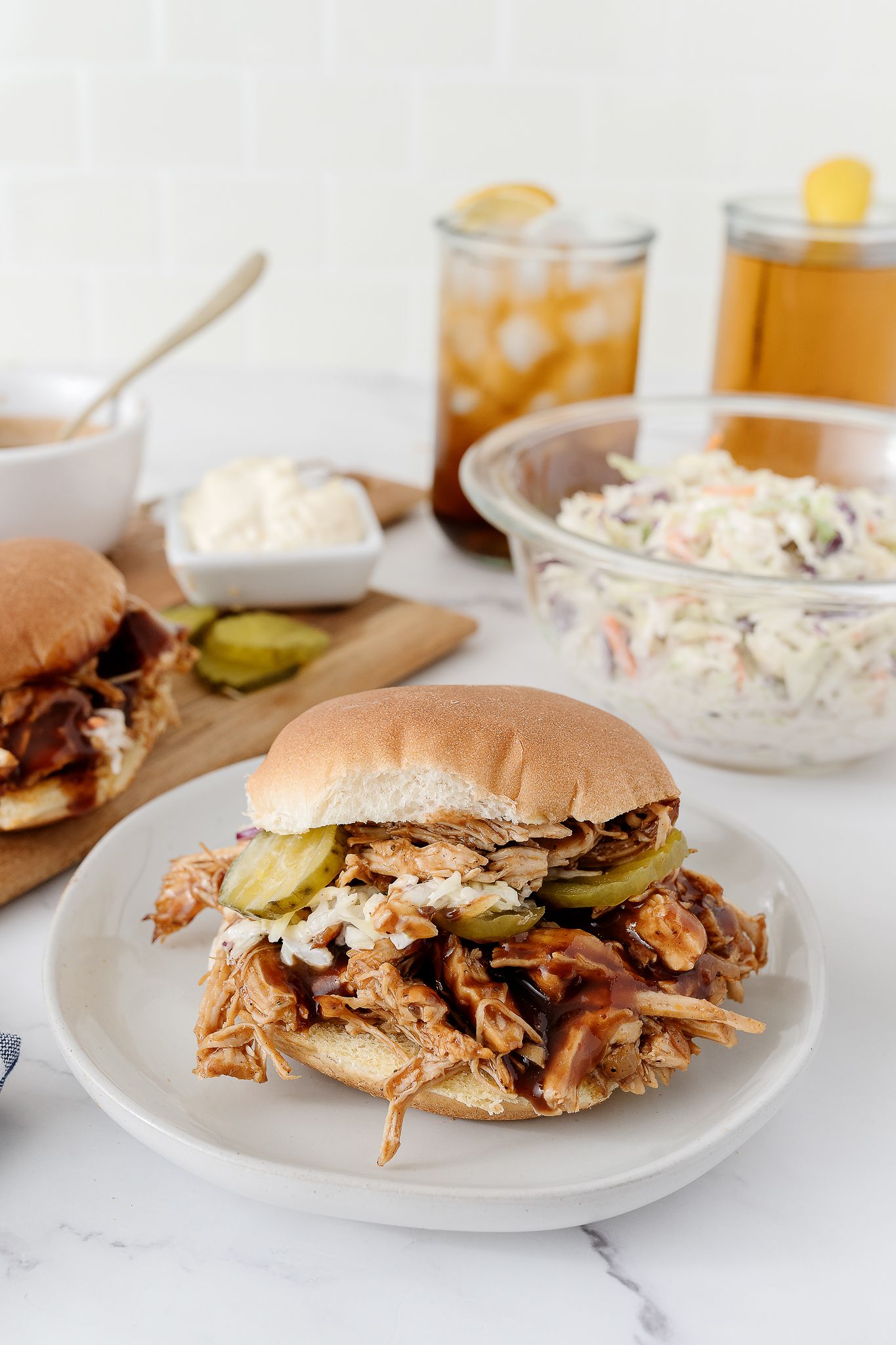 Pulled pork sandwich with pickles on a white plate served with coleslaw and iced tea.