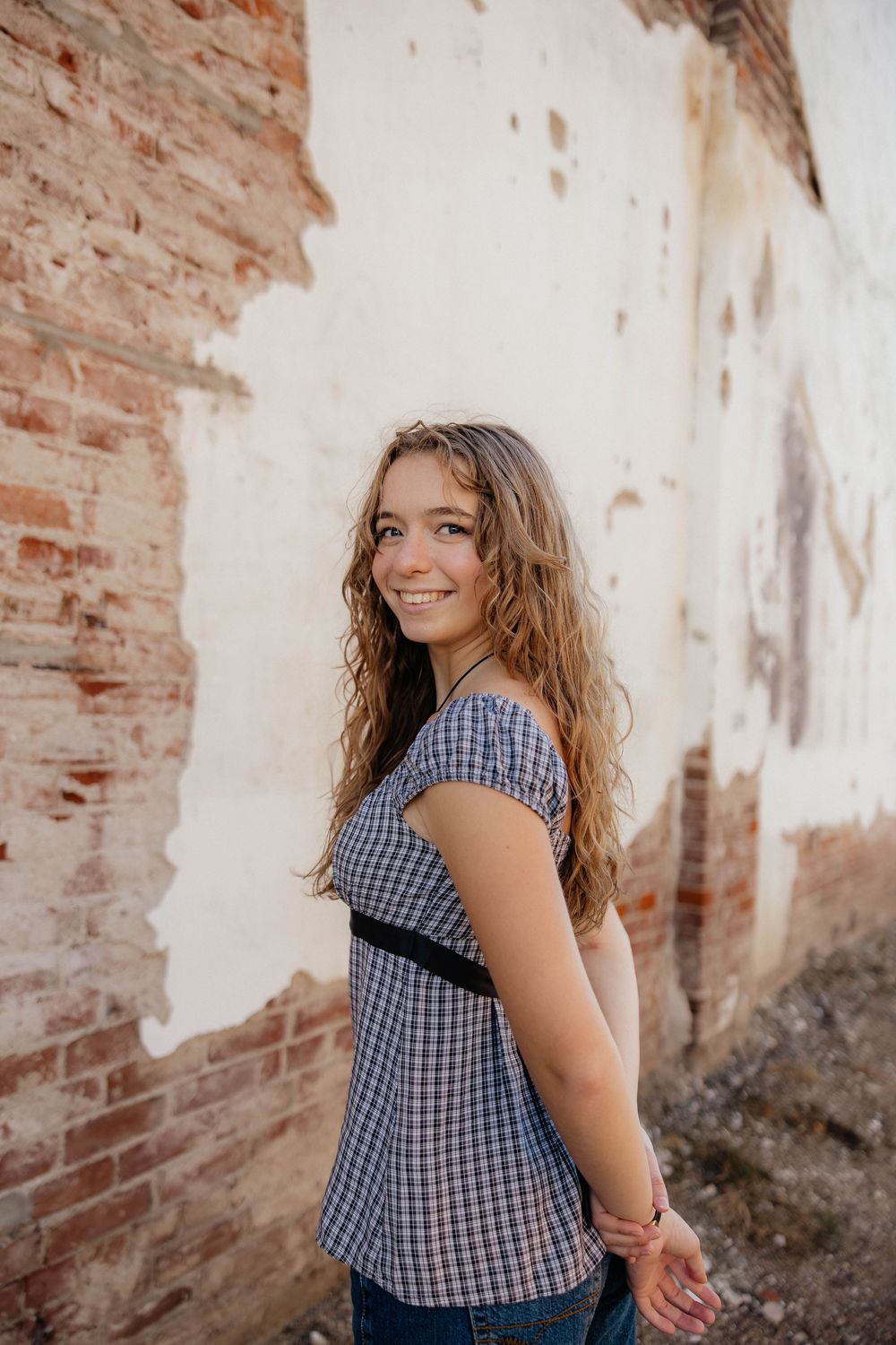 A person in a blue gingham top poses against a rustic brick wall on a sunny day.