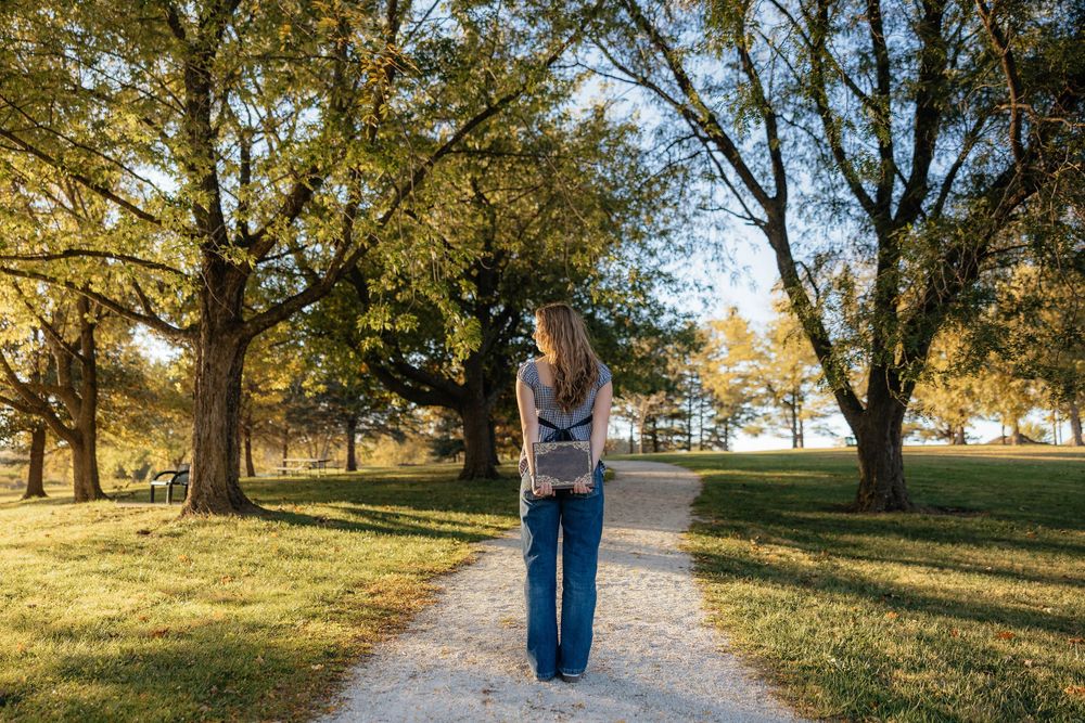 A gravel walking path winds through a scenic park with golden autumn trees casting long shadows in warm sunlight.