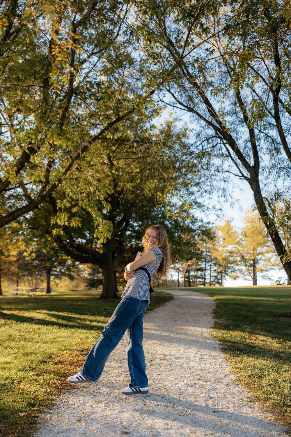 Person in jeans walks along a sunlit path through a park with golden autumn trees casting shadows.