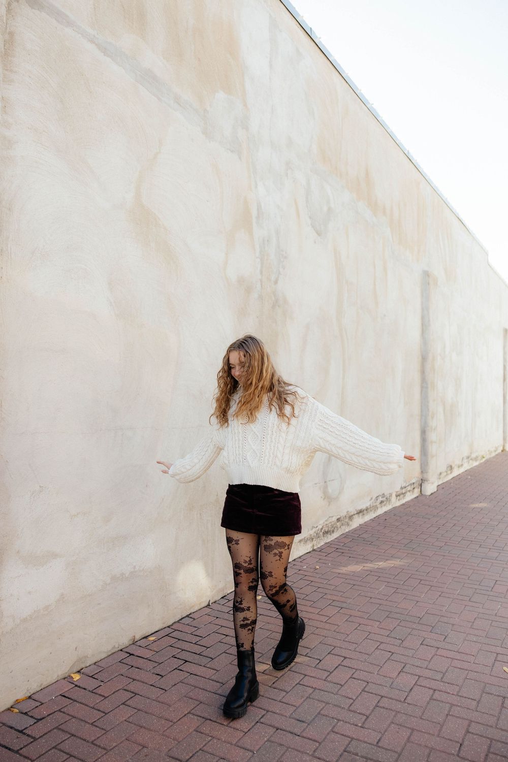 A person in a white sweater and black skirt with patterned tights poses playfully against a beige wall.