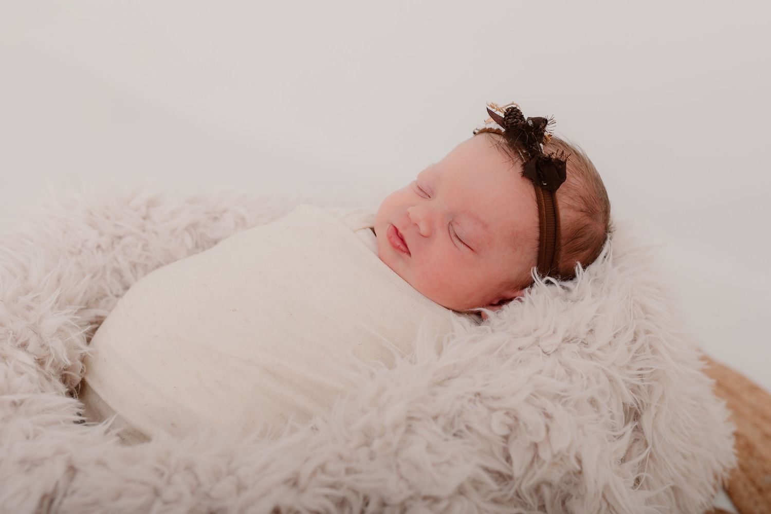 Sleeping newborn wrapped in white swaddle with brown headband on fluffy blanket.