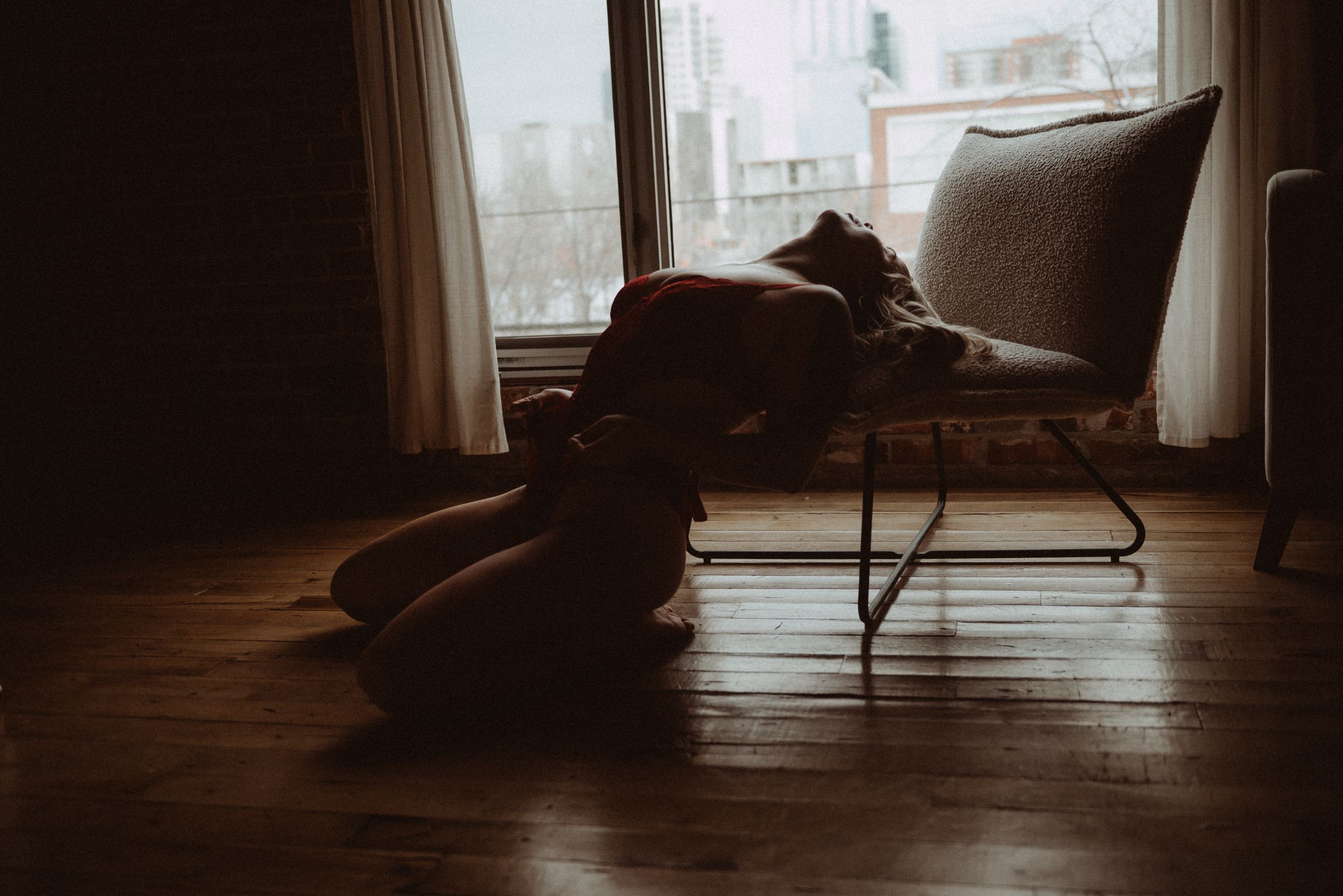 A silhouetted figure rests against an armchair near a window in a dimly lit room with hardwood floors.