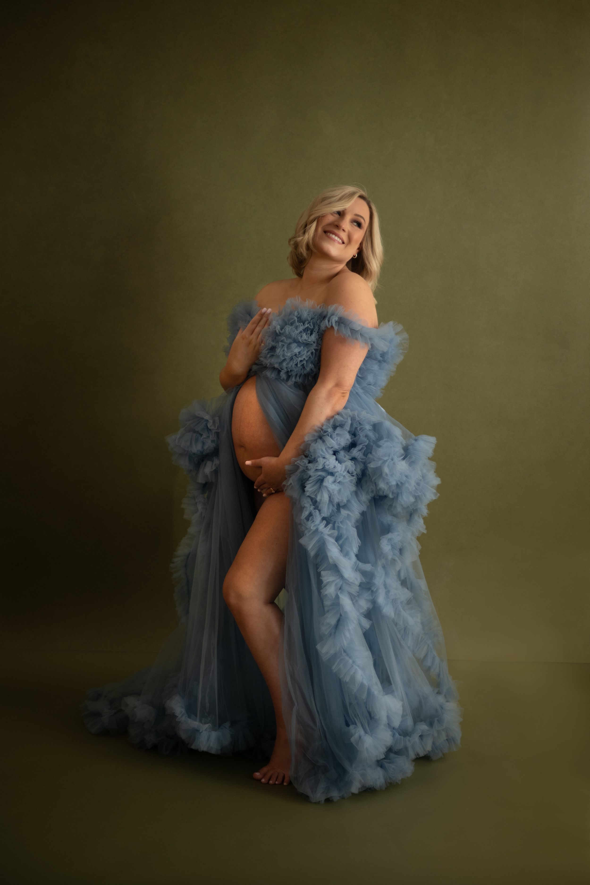 A person in a flowing dusty blue tulle gown poses gracefully against a dark olive backdrop in a maternity photo session.