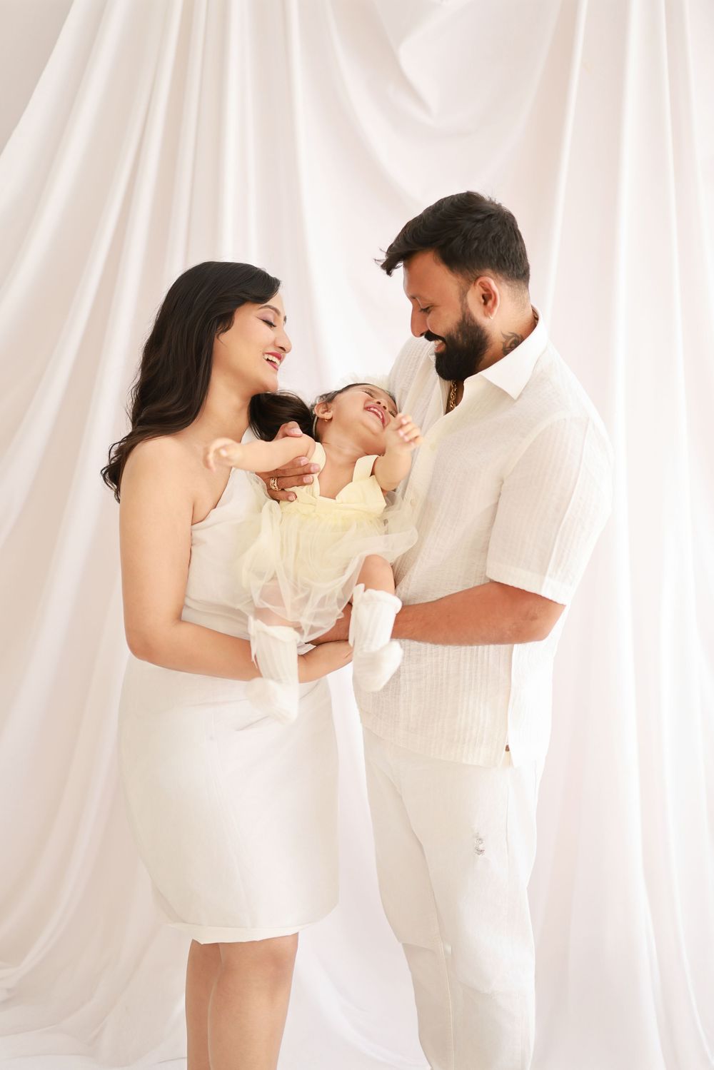 A couple in white outfits shares intimate moments in a bright studio setting with flowing curtains.