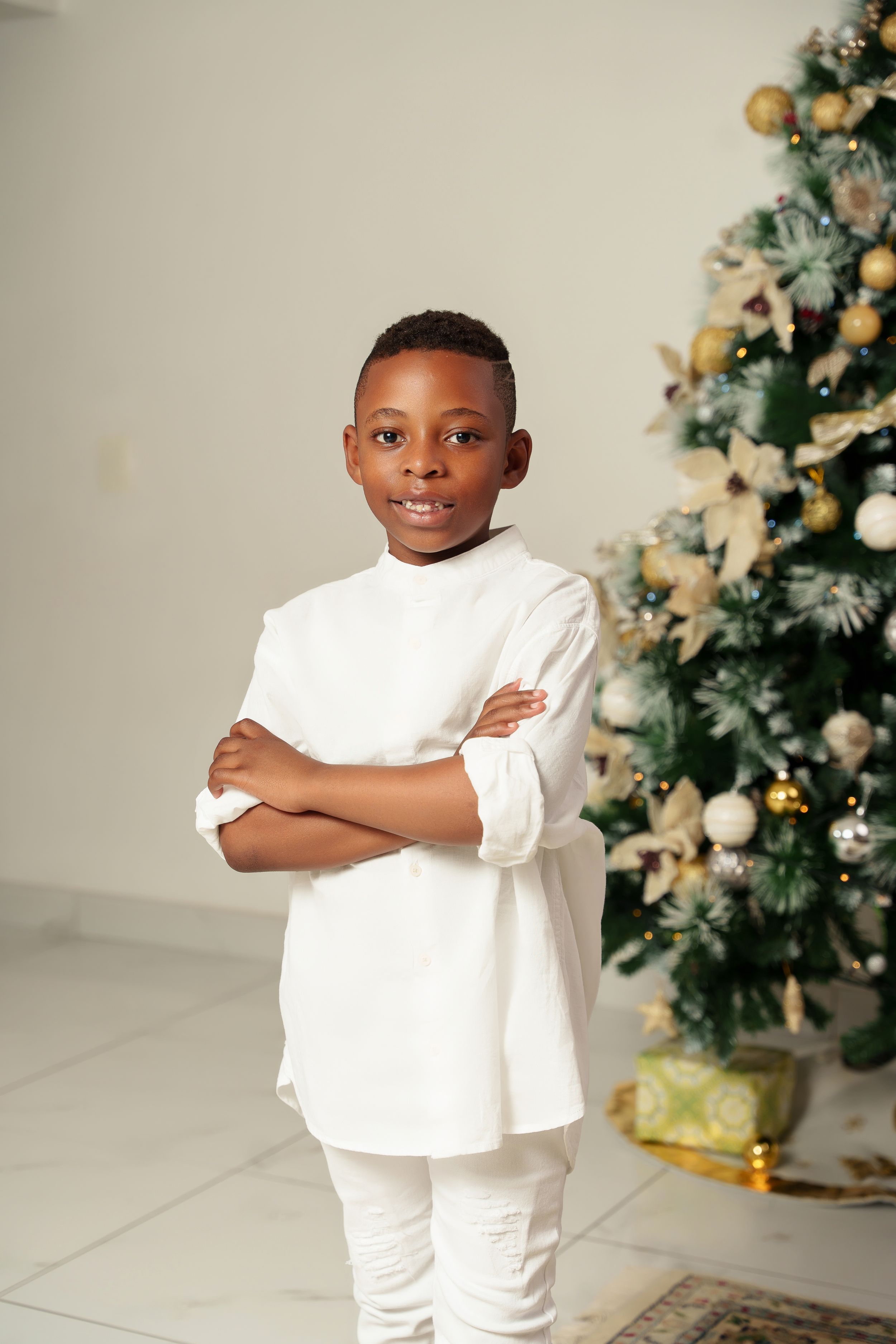 Young person in all white outfit poses confidently in front of Christmas tree.