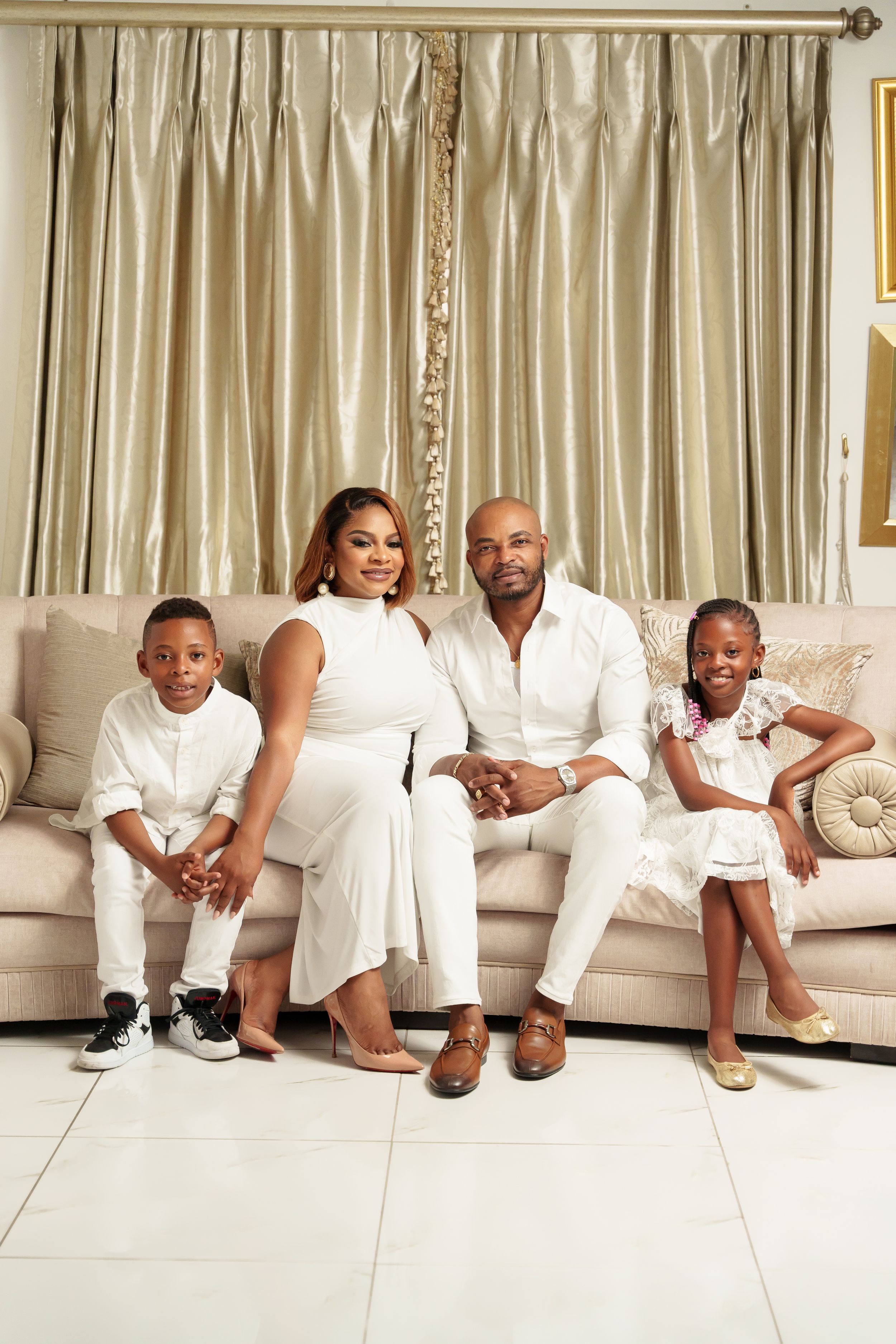 Family dressed in white sits together on beige couch for formal portrait.