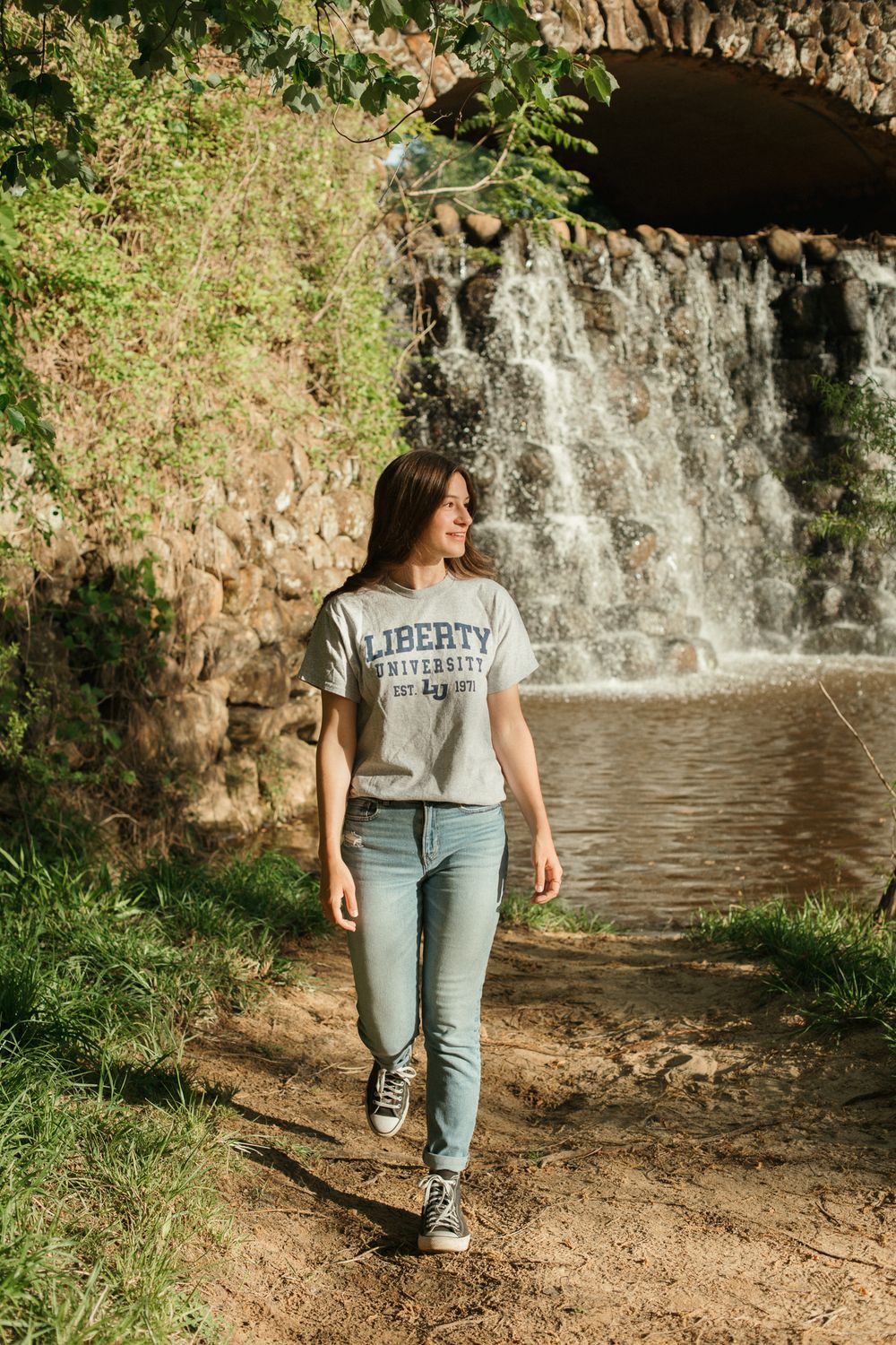 Student in Liberty University gear walks along scenic waterfall path.